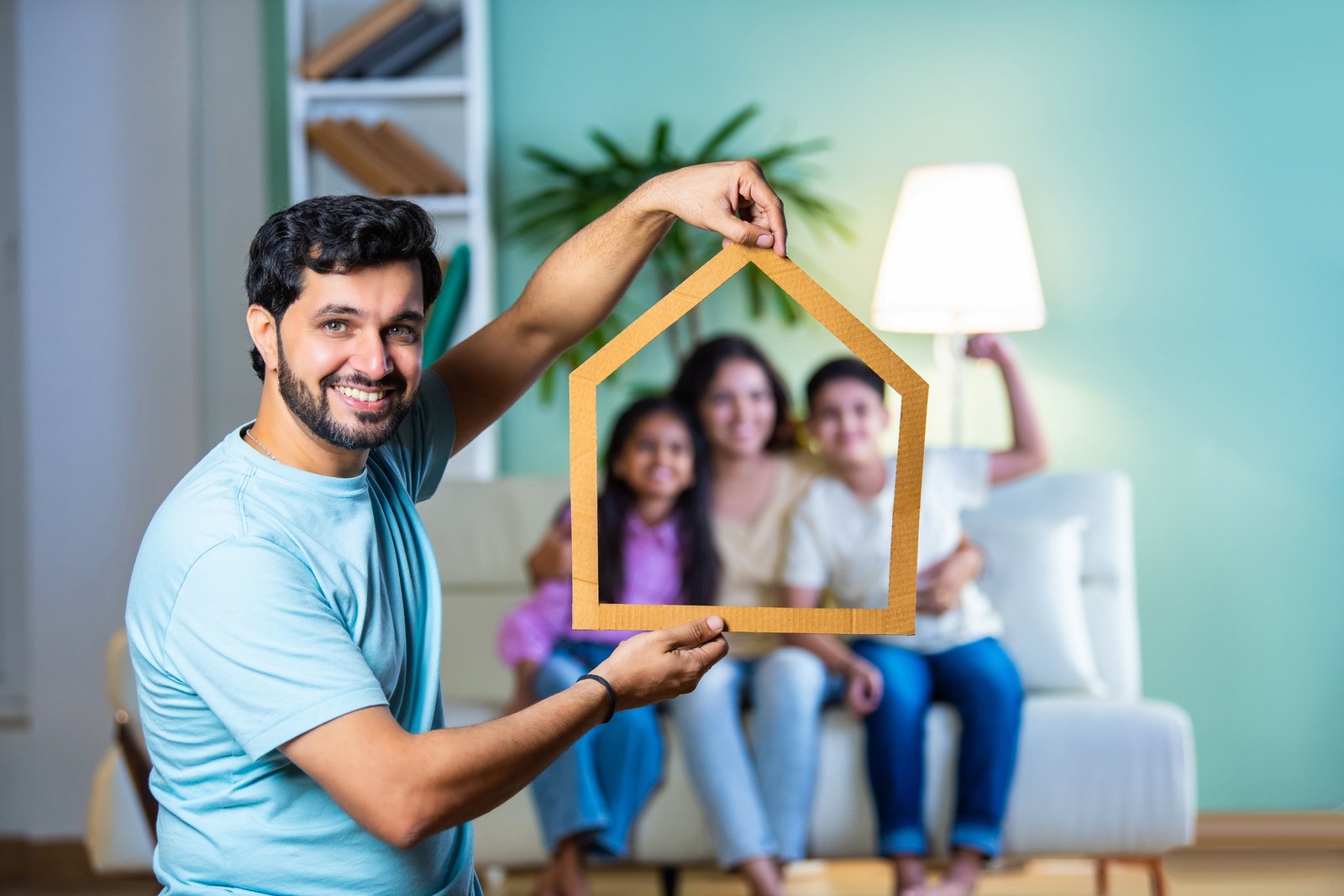 Indian dad holding a cardboard home cutout as his family sits on the sofa, real estate dreams